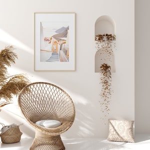 May include: A white wicker chair with a white cushion sits in a room with white walls. A framed photograph of a white building with a staircase hangs on the wall. A brown plant hangs from a niche in the wall.