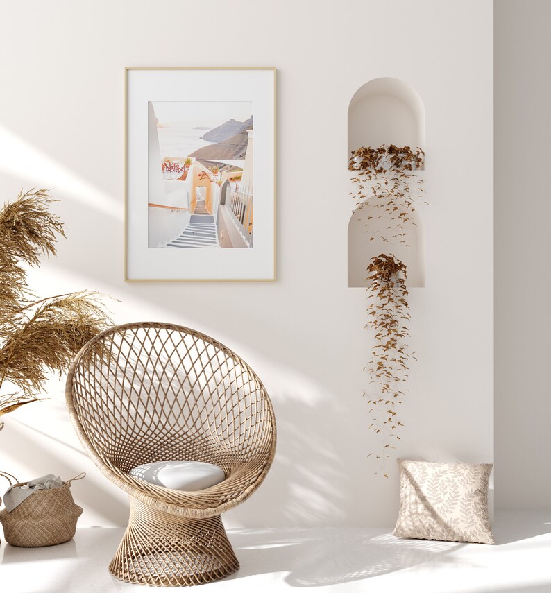 May include: A white wicker chair with a white cushion sits in a room with white walls. A framed photograph of a white building with a staircase hangs on the wall. A brown plant hangs from a niche in the wall.