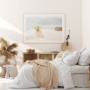May include: A white bed with a tan throw blanket and white pillows. The bed is in a bedroom with a white wall and a framed photograph of a landscape with a plant in a pot. There is a wooden cabinet with glass doors behind the bed.