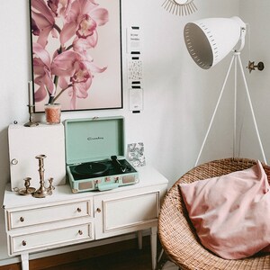 May include: A white dresser with three drawers and gold knobs, a green vintage record player, a pink orchid print, a wicker chair with a pink pillow, and a white floor lamp with a white shade.