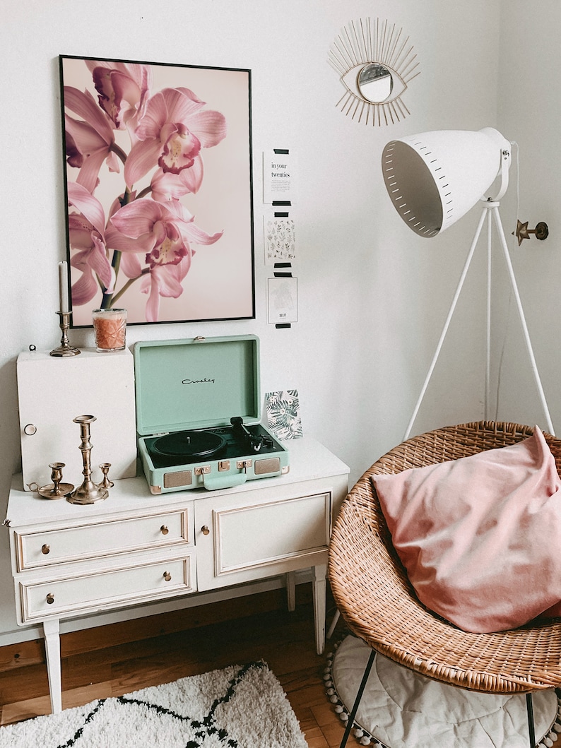 May include: A white dresser with three drawers and gold knobs, a green vintage record player, a pink orchid print, a wicker chair with a pink pillow, and a white floor lamp with a white shade.
