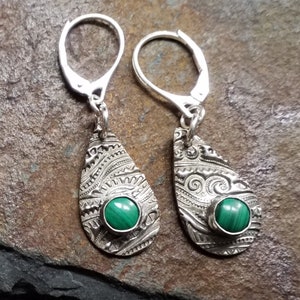 May include: A pair of silver earrings with a green malachite stone set in each. The earrings have a teardrop shape and are decorated with intricate patterns.