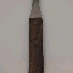May include: A two-pronged carving fork featuring a metallic silver head and a dark brown wooden handle. The handle is secured with two circular rivets and displays a small, faint engraved bull head logo near the bottom edge of the wood.