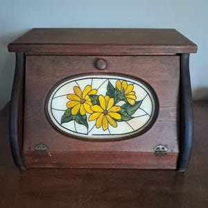 May include: A dark brown wooden bread box with a drop-down front door featuring a central oval glass panel. The glass displays a painted design of three yellow flowers with green leaves and black outlines. The door has a small round wooden knob.