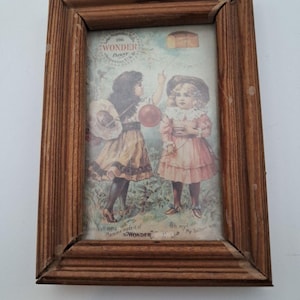 May include: A framed vintage print featuring two young girls in dresses, one holding a red balloon. The print is in a wooden frame. The print has text that reads "1906 Wonder Patent Superior Mills U.S."