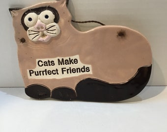 May include: A ceramic cat-shaped wall hanging in shades of pink and brown. The cat has large, expressive eyes, a pink nose, and a white mouth. The text "Cats Make Purrfect Friends" is displayed on the cat's body. A brown string is attached for hanging.