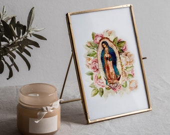 May include: A gold-framed picture with a white background and a colorful image of the Virgin Mary surrounded by pink roses. The picture is on a white linen table cloth with a clear glass vase with a green plant and a brown candle.