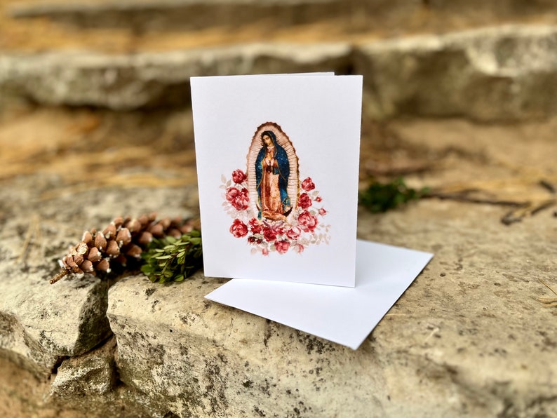 Op de afbeelding: Een witte kaart met een kleurrijk beeld van de Maagd Maria, omringd door rode rozen. Het beeld is in een ovale lijst.