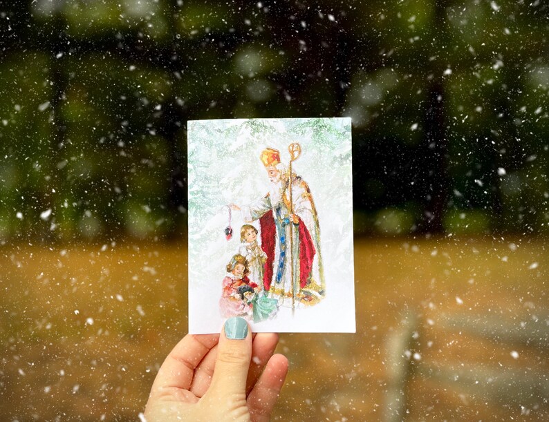 Op de afbeelding: Een kerstkaart met een afbeelding van Sinterklaas met twee kinderen. Sinterklaas draagt een rode en gouden mantel en een mijter. De kinderen zijn feestelijk gekleed. De kaart heeft een besneeuwde achtergrond.