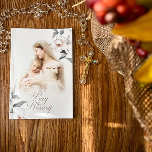 Könnte beinhalten: Ein weißes Büchlein mit den Worten "Pray the Rosary" auf dem Umschlag. Der Umschlag zeigt ein Gemälde von Maria, die das Jesuskind und ein weißes Lamm hält. Eine goldene Rosenkranz mit einem Kristallkreuz liegt auf einer Holzoberfläche.
