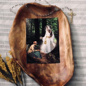 Puede incluir: Un cuenco de madera de forma orgánica contiene una impresión enmarcada de una escena religiosa. La impresión representa a una mujer con un vestido blanco con un halo, interactuando con otra mujer en un entorno forestal. Un rosario y una cruz están sobre el cuenco.
