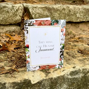 Op de afbeelding: Een kerstkaart met een bloemmotief en gouden accenten. De kaart bevat de tekst "Zij zullen Hem Immanuel noemen." en de bijbelverwijzing "Matthe&uuml;s 1:23."