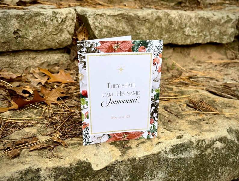 Op de afbeelding: Een kerstkaart met een bloemmotief en gouden accenten. De kaart bevat de tekst "Zij zullen Hem Immanuel noemen." en de bijbelverwijzing "Matthe&uuml;s 1:23."