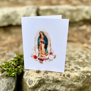 Op de afbeelding: Een witte kaart met een aquarelillustratie van Onze Lieve Vrouw van Guadalupe. De afbeelding toont haar staand in een blauwe en gouden jurk met een kroon van sterren. Ze is omringd door witte bloemen.