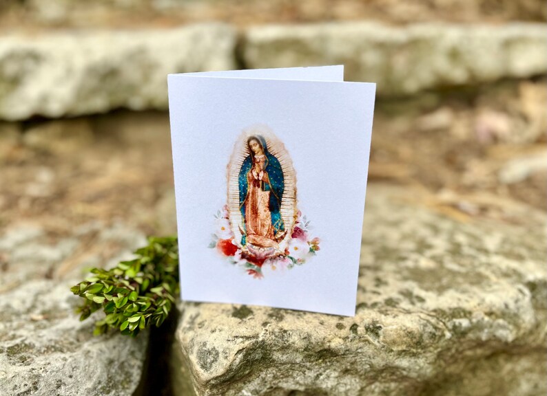 Op de afbeelding: Een witte kaart met een aquarelillustratie van Onze Lieve Vrouw van Guadalupe. De afbeelding toont haar staand in een blauwe en gouden jurk met een kroon van sterren. Ze is omringd door witte bloemen.
