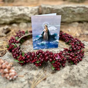 Op de afbeelding: Een kaart met een religieus beeld van een vrouw in een blauwe en witte jurk die op water staat. De kaart is omringd door een krans van rode bessen en groene bladeren.