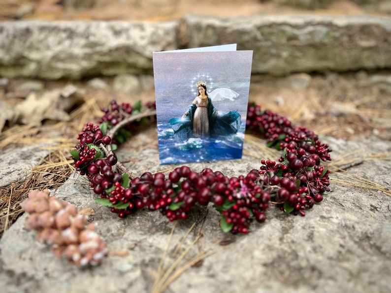 Op de afbeelding: Een kaart met een religieus beeld van een vrouw in een blauwe en witte jurk die op water staat. De kaart is omringd door een krans van rode bessen en groene bladeren.