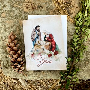Op de afbeelding: Een kerstkaart met een aquarel van de kerststal met Maria, Jozef en het kindje Jezus. De kaart heeft een witte achtergrond met een vintage textuur en het woord "Gloria" in rode cursieve letters.