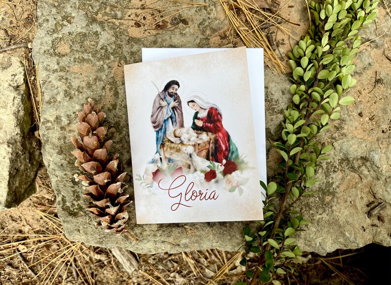 Op de afbeelding: Een kerstkaart met een aquarel van de kerststal met Maria, Jozef en het kindje Jezus. De kaart heeft een witte achtergrond met een vintage textuur en het woord "Gloria" in rode cursieve letters.