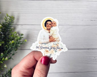 May include: A sticker featuring a watercolor illustration of Saint Gianna with a baby in her arms. The sticker has a gold halo around Saint Gianna's head and the text "St. Gianna, pray for us" at the bottom.