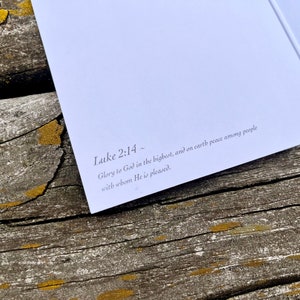 Op de afbeelding: Een close-up van een witte kaart met de tekst "Lukas 2:14~ Ere zij God in de hoogste, en vrede op aarde onder de mensen die Hij welgevallig zijn." gedrukt in zwarte inkt.