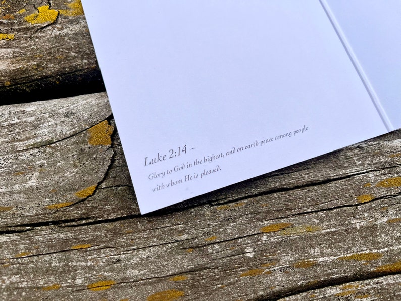 Op de afbeelding: Een close-up van een witte kaart met de tekst "Lukas 2:14~ Ere zij God in de hoogste, en vrede op aarde onder de mensen die Hij welgevallig zijn." gedrukt in zwarte inkt.
