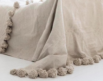 May include: A beige throw blanket with a fringe of pom-poms around the edges. The blanket is draped over a bed.