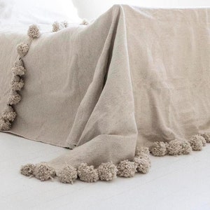 May include: A beige throw blanket with a fringe of pom-poms around the edges. The blanket is draped over a bed.