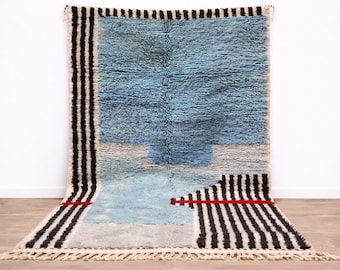 Puede incluir: Una alfombra rectangular con un diseño azul y blanco. La alfombra tiene un campo azul central con una sección azul claro y gris en la parte inferior. Los bordes y acentos a rayas blancas y negras, con una raya roja, enmarcan la alfombra. La alfombra tiene un borde con flecos.
