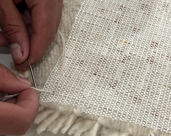Könnte beinhalten: Nahaufnahme der Teppichreparatur. Eine Hand verwendet Nadel und Faden, um die Rückseite eines cremefarbenen Teppichs zu nähen. Der Teppich hat eine weiße Netzrückseite und einen Fransen aus weißem Garn. Der Teppich hat ein beige-braunes Muster.