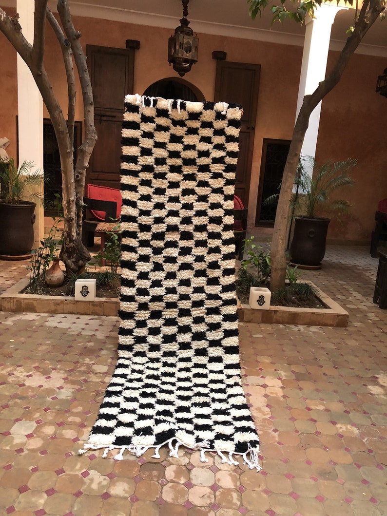 May include: A black and white checkered rug with a fringe border. The rug is made of a soft, fluffy material and is hanging on a wall in a courtyard.