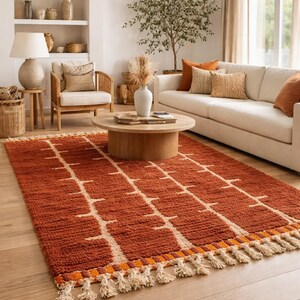 May include: A rectangular rust-colored rug with a geometric pattern of cream-colored lines and tassels along the edge. The rug is placed on a hardwood floor in a living room setting, with a sofa and coffee table.