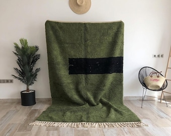 Op de afbeelding: Een groot, rechthoekig, olijfgroen vloerkleed met een dikke, horizontale zwarte streep in het midden. Het kleed heeft een diepe, hoogpolige textuur en is afgewerkt met lange, crèmekleurige franjes aan de onderkant.
