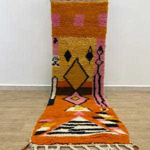Puede incluir: Una vibrante alfombra rectangular con un diseño geométrico. La alfombra presenta una combinación de colores naranja, rosa, amarillo, negro y blanco. El diseño incluye formas de diamante, rayas y otros patrones abstractos. La alfombra tiene un borde con flecos.