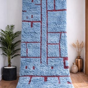 May include: A large, rectangular blue rug with a geometric burgundy design. The rug has a shaggy texture and a cream-colored fringe along the bottom edge. The rug is displayed against a white wall, with a potted plant and decorative vases in the background.