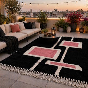 K&ouml;nnte beinhalten: Ein schwarzer Teppich mit geometrischem Muster in Wei&szlig; und Rosa, mit Fransen. Der Teppich befindet sich auf einer Terrasse mit Sofa, Pflanzen und Lichterketten, wodurch ein gem&uuml;tlicher Au&szlig;enbereich entsteht.