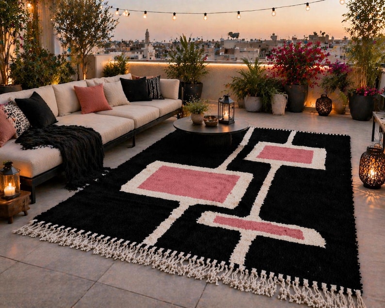 K&ouml;nnte beinhalten: Ein schwarzer Teppich mit geometrischem Muster in Wei&szlig; und Rosa, mit Fransen. Der Teppich befindet sich auf einer Terrasse mit Sofa, Pflanzen und Lichterketten, wodurch ein gem&uuml;tlicher Au&szlig;enbereich entsteht.