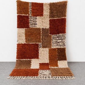Peut inclure: Un tapis rectangulaire et touffu avec un motif patchwork. Le tapis présente différentes nuances de marron, de beige et de carrés de couleur rouille. Le bord inférieur a une frange. Le tapis est présenté contre un mur blanc et un sol gris.