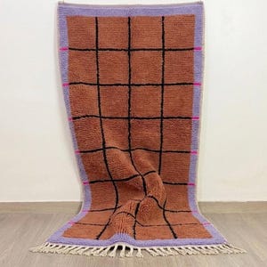 Peut inclure: Un tapis rectangulaire présentant un champ brun terre cuite texturé avec un motif de grille noir. Le tapis est bordé d'un cadre violet clair accentué de petits tirets horizontaux roses sur les côtés et fini avec des franges couleur crème.