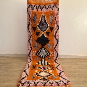 May include: A vibrant orange and pink Moroccan rug featuring a central diamond design in black, white, and grey. The rug has a black and pink zig-zag border. The rug is made of a thick, textured material.