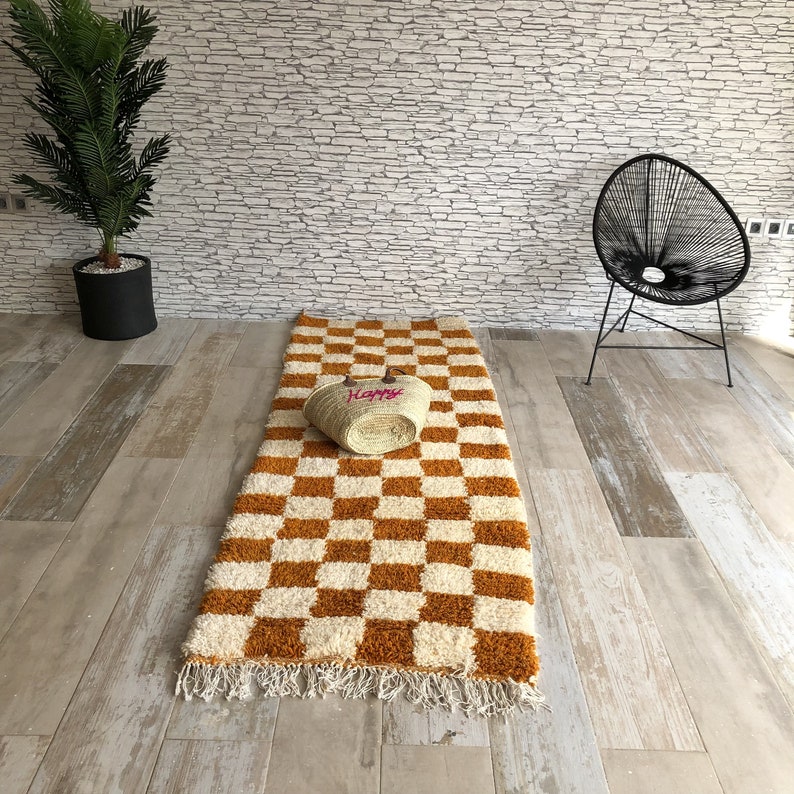 May include: A checkered rug with white and orange squares. The rug has a fringe border and a straw basket with the word "Happy" embroidered on it is sitting on the rug.