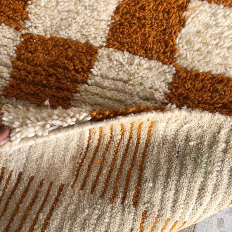 May include: Close-up of a white and orange woven rug with a geometric pattern. The rug is folded in half, showing the underside with a striped pattern.