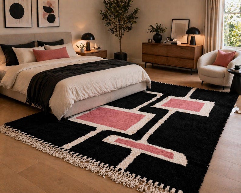 K&ouml;nnte beinhalten: Ein rechteckiger Teppich mit schwarzem Hintergrund und einem geometrischen Muster aus wei&szlig;en Linien und rosafarbenen rechteckigen Bl&ouml;cken. Der Teppich hat einen dichten, strukturierten Flor und ist an den kurzen Seiten mit langen, geknoteten Fransen versehen.