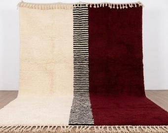Op de afbeelding: Een rechthoekig tapijt met een colorblock-ontwerp. De ene kant is crèmekleurig, de andere is diep bordeauxrood. Een verticale streep van zwarte en witte lijnen verdeelt de twee kleuren. Het tapijt heeft franjes aan de boven- en onderkant.