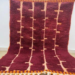May include: A burgundy and cream-colored rug with a geometric pattern. The rug features a repeating design of vertical lines with horizontal blocks, creating a textured appearance. The edges are trimmed with orange and cream fringe.