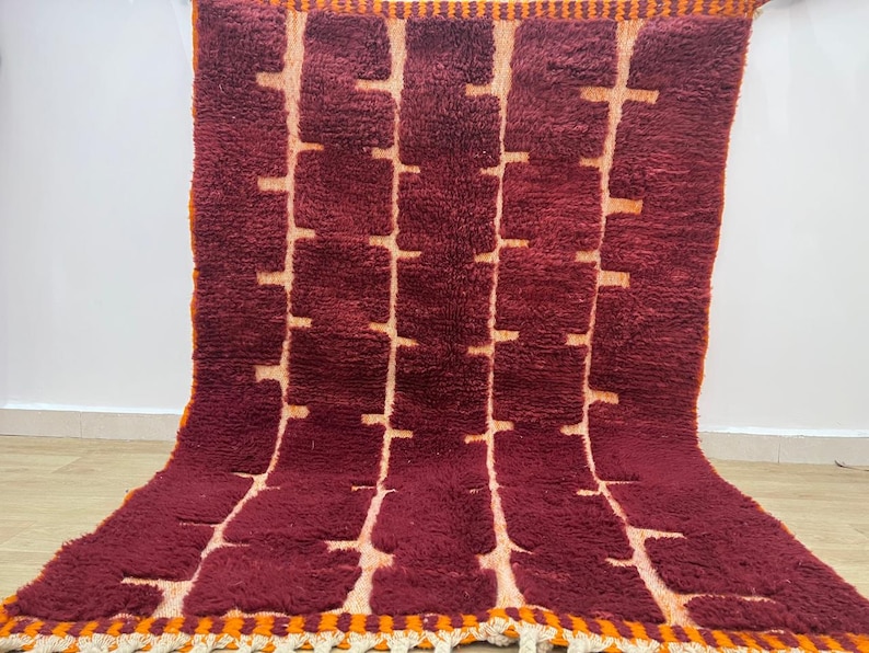 May include: A burgundy and cream-colored rug with a geometric pattern. The rug features a repeating design of vertical lines with horizontal blocks, creating a textured appearance. The edges are trimmed with orange and cream fringe.