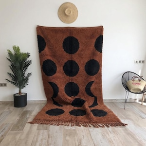 Peut inclure: Un tapis rectangulaire à poils longs de couleur rouille, orné d'un motif répétitif de grands cercles noirs pleins et de deux formes de croissant de lune noires. Le tapis présente un poil épais et texturé avec une frange de pompons en bas.