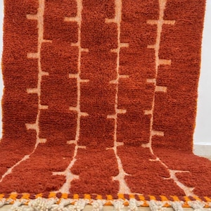 May include: A rust-colored, textured rug with a geometric pattern of vertical lines and cross-like shapes in a lighter shade. The rug has a fringe along the bottom edge. The rug is made of a thick, plush material.