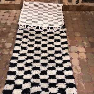 May include: Two black and white patterned rugs with fringe. The top rug has a striped pattern and the bottom rug has a checkerboard pattern.