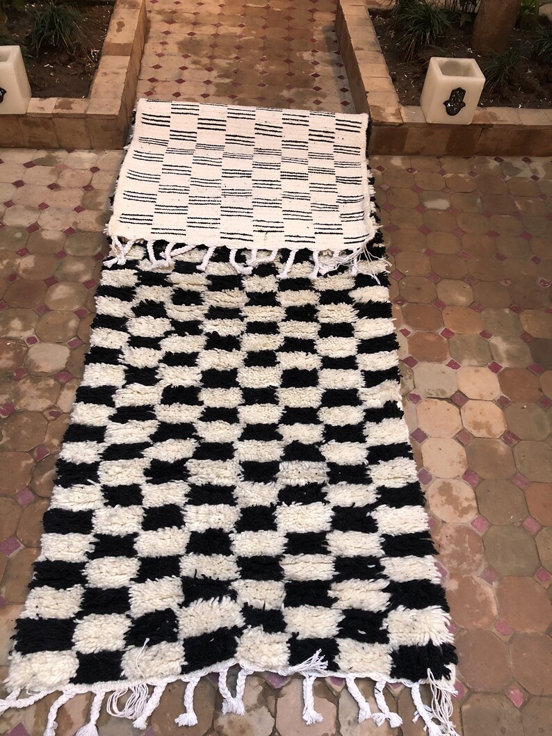 May include: Two black and white patterned rugs with fringe. The top rug has a striped pattern and the bottom rug has a checkerboard pattern.